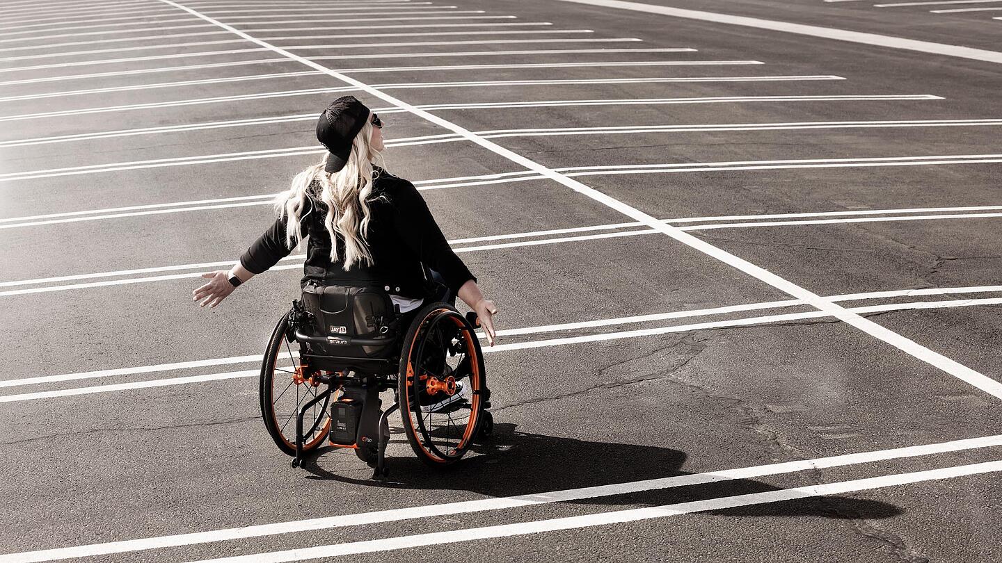 Frau im Rollstuhl auf leerem Parkplatz, Bildquelle: 2025, Sunrise Medical GmbH, Malsch/HD Frau im Rollstuhl steht auf leerem Parkplatz und hält die Arme zur Seite, Bildquelle: 2025, Sunrise Medical GmbH, Malsch/HD