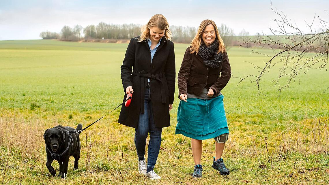 Hundespaziergang über eine Wiese mit Fußhebe-Schwäche, © Ottobock zwei Frauen die mit einem Hund über eine Wiese spazieren, eine trägt eine Knie-Manschette aufgrund einer Fußhebe-Schwäche, © Ottobock