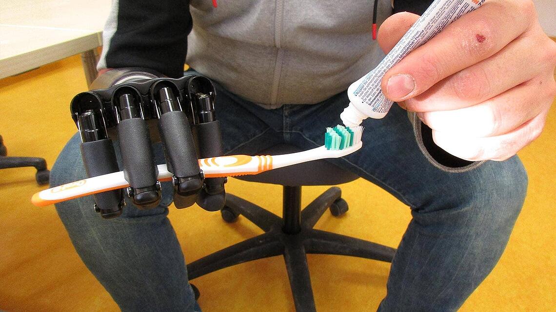 Zahnpasta auf Zahnbürste geben bei Prothesen-Gebrauchsschulung Mann sitzt auf Stuhl und hat an rechter Hand Prothese, mit linker Hand gibt er Zahnpasta auf die Zahnbürste bei der Prothesen-Gebrauchsschulung