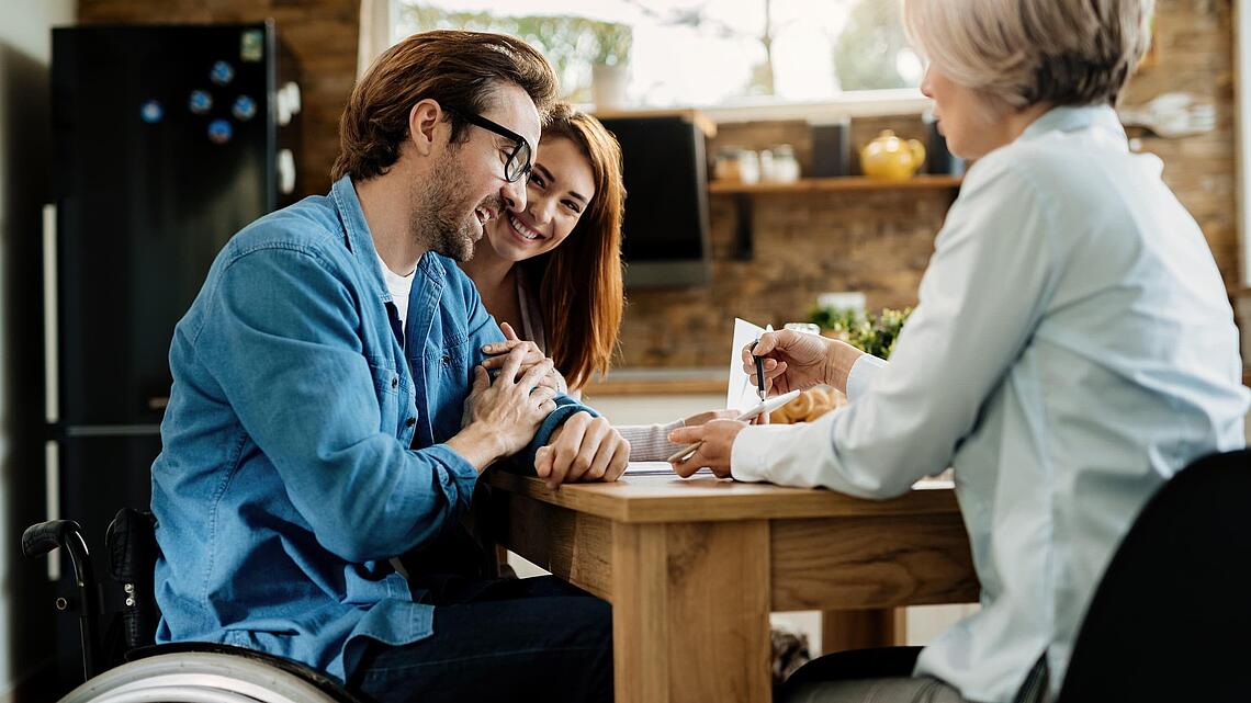 Junger Mann wird an einem Holztisch von einer Frau zu seinem neuen Rollstuhl beraten; im Hintegrund schaut die Frau des Rollstuhlfahrers glücklich zu