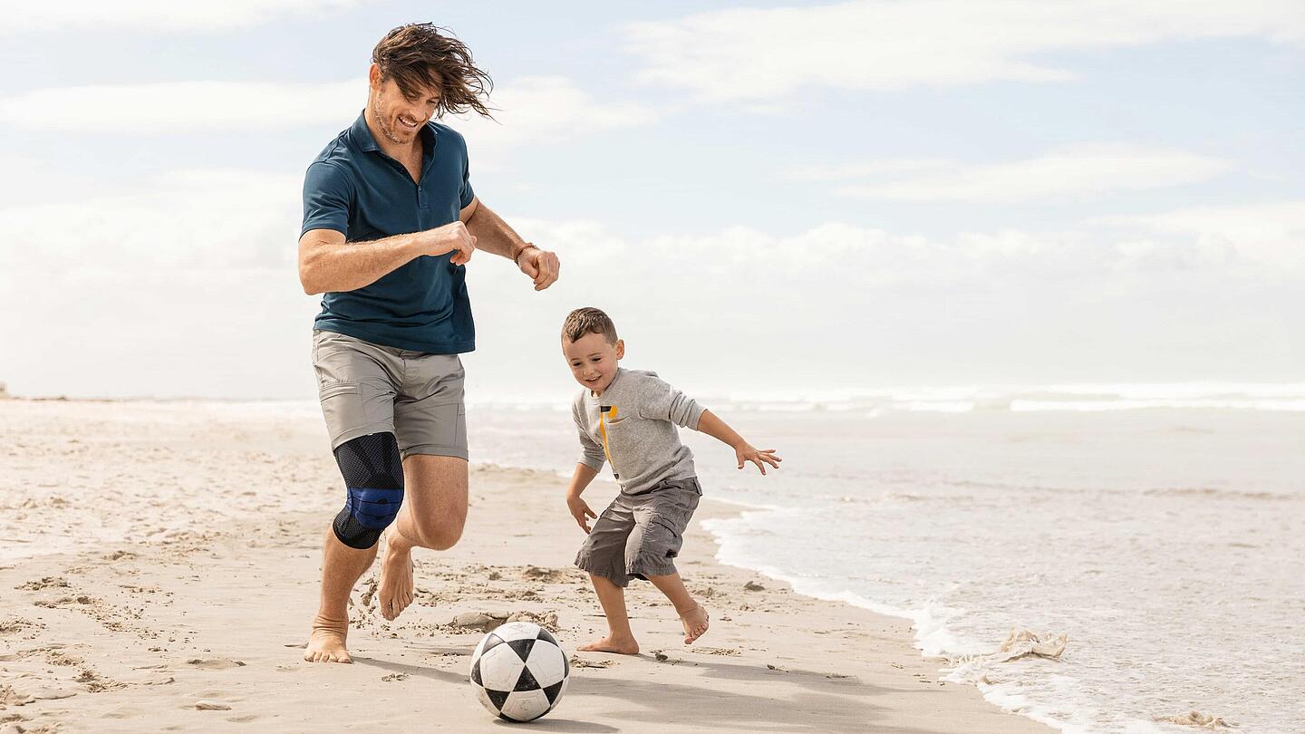 Fußballspielen mit Kniebandage Genutrain, Bild: Bauerfeind Mann mit schwarzer Kniebandage Genutrain spielt Fußball mit Kind am Strand, Bild: Bauerfeind